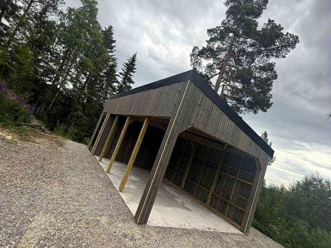 En åpen carport i mørkt tre på en grusplass i skogen