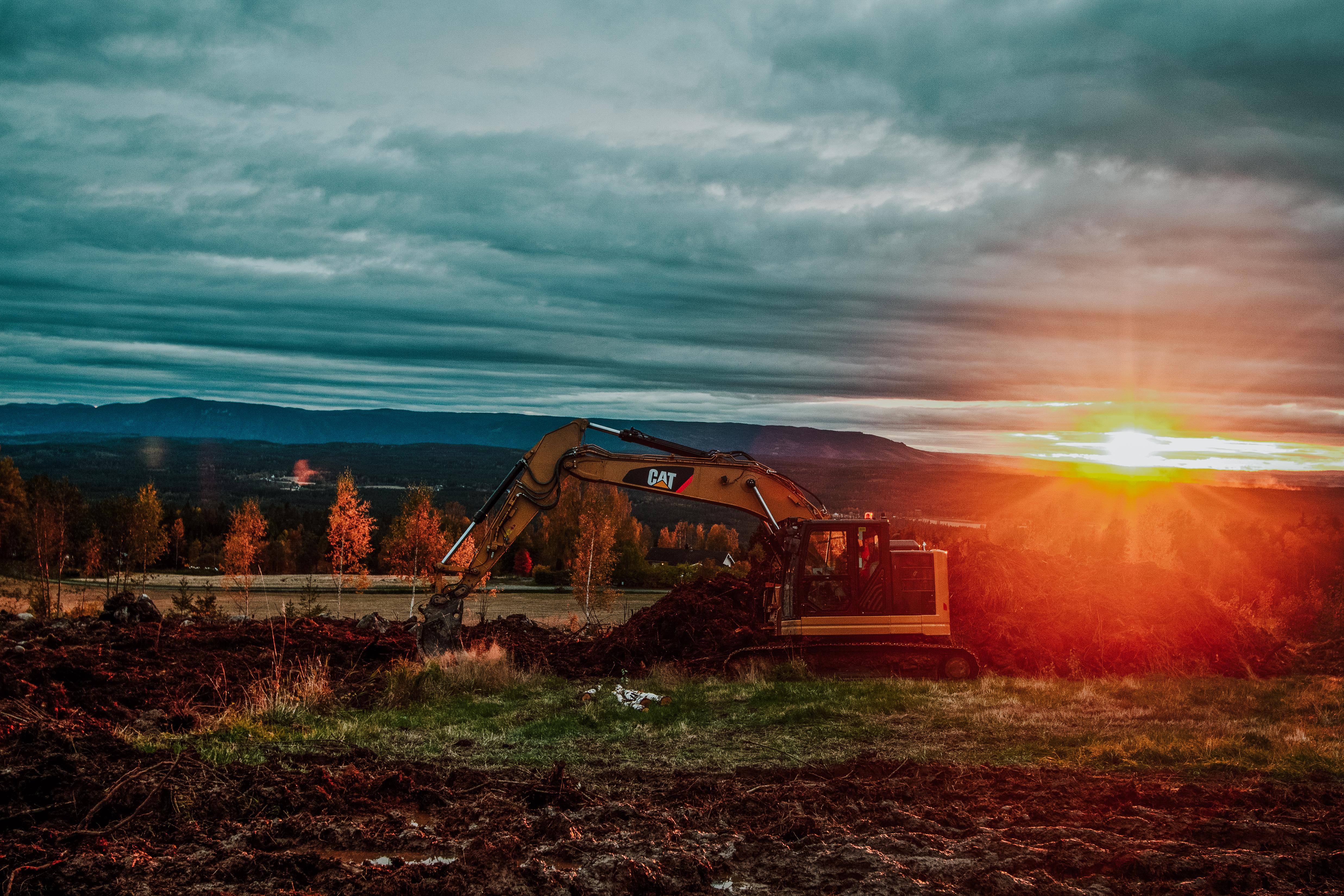 En gravemaskin på et jorde i solnedgang, med skog og åser bak